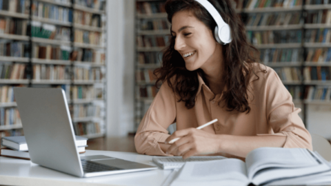 The woman in front of the laptop, wearing earphones, holding a pencil and notebook, and watching the HeartCount webinar on Happiness helps any business thrive